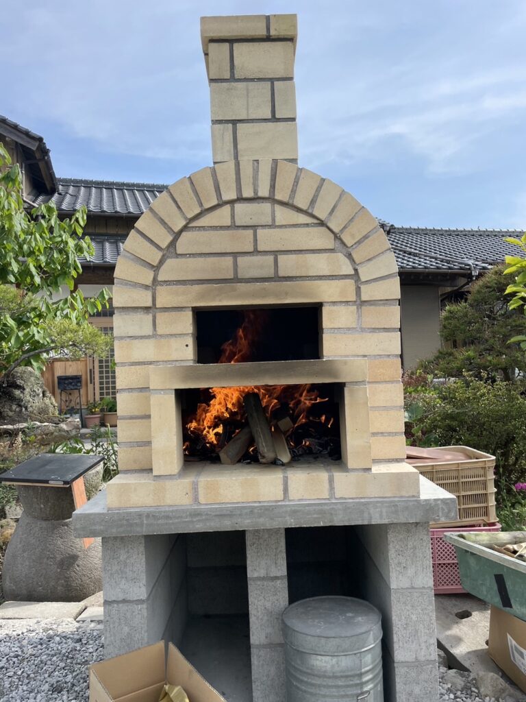 完成したばかりのモダンなデザインの庭用ピザ窯。炎が燃える窯の内部と、焼きあがったばかりのピザ。リフォームによる外構の変化。