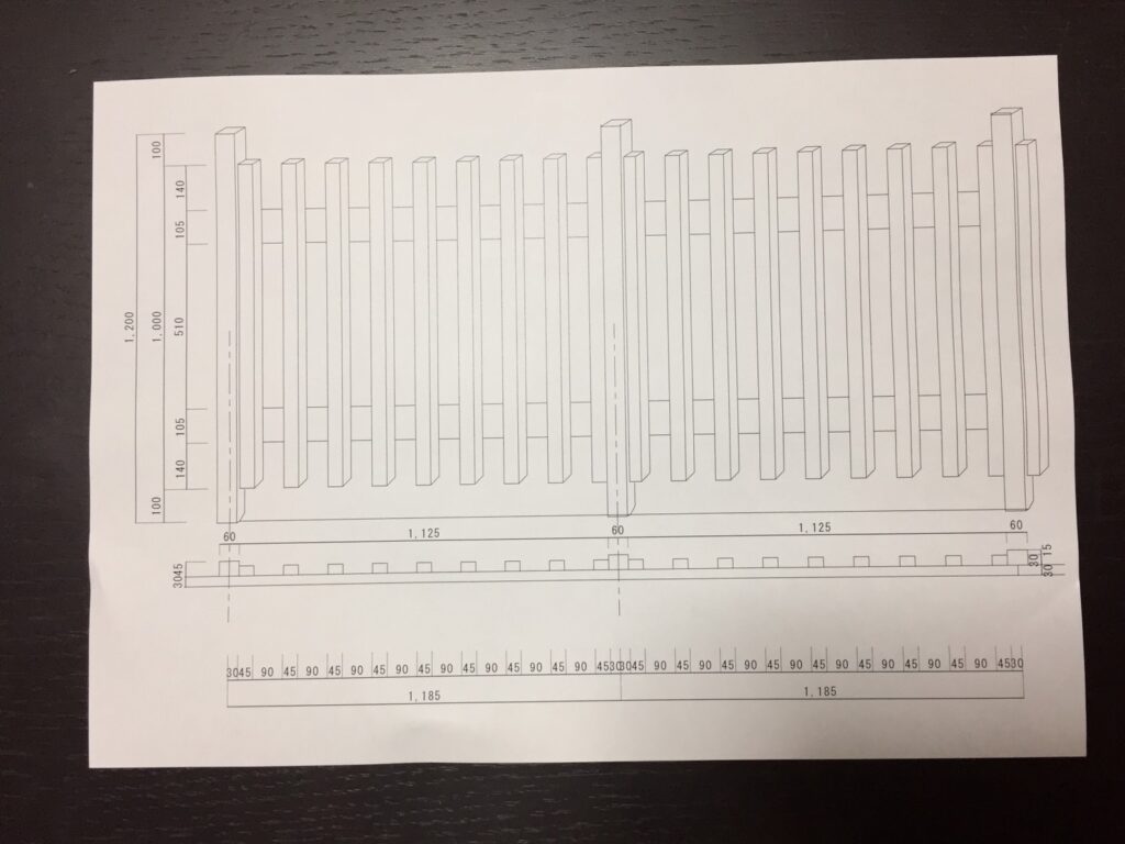 建築会社のスタッフが、自宅のDIY木製フェンスの設計図(手書きのスケッチや寸法が描かれた図面)を見ながら計画している様子。テーブルには鉛筆や定規が置かれ、具体的な作業工程を考えている。