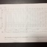 建築会社のスタッフが、自宅のDIY木製フェンスの設計図（手書きのスケッチや寸法が描かれた図面）を見ながら計画している様子。テーブルには鉛筆や定規が置かれ、具体的な作業工程を考えている。