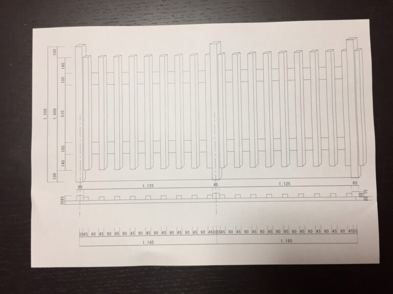 建築会社のスタッフが、自宅のDIY木製フェンスの設計図（手書きのスケッチや寸法が描かれた図面）を見ながら計画している様子。テーブルには鉛筆や定規が置かれ、具体的な作業工程を考えている。
