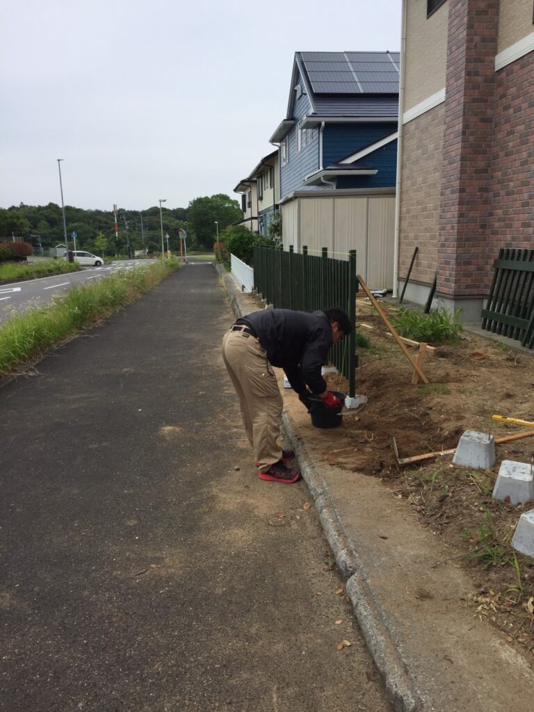日本の庭で、男性がDIYで木製縦格子フェンスを設置している作業中の写真。フェンスを基礎に固定し、ドリルや水平器などの工具が周囲に置かれ、作業の様子がうかがえる。