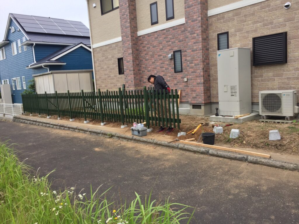 外で、男性がDIYで木製縦格子フェンスを設置している作業中の写真。フェンスを基礎に固定し、ドリルや水平器などの工具が周囲に置かれ、作業の様子がうかがえる。