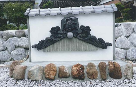白壁と瓦で仕上げた和風モニュメントの中央に、3つのパーツで構成された鬼瓦が据えられた完成写真。伝統的な雰囲気の中に力強い存在感がある。