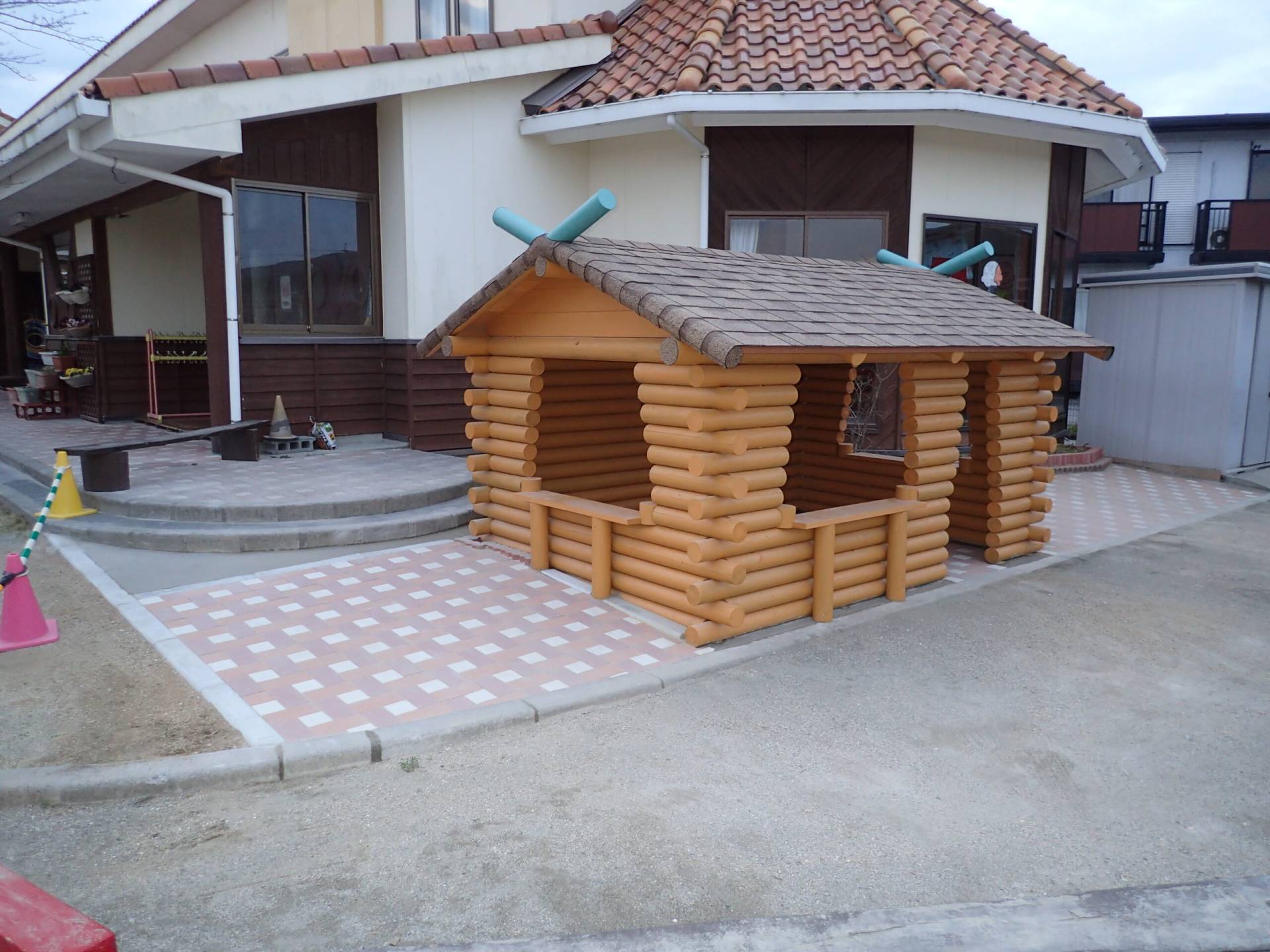 インターロッキングの床とシングル葺きの屋根を持つ、こども園の野外遊具としての丸太小屋。