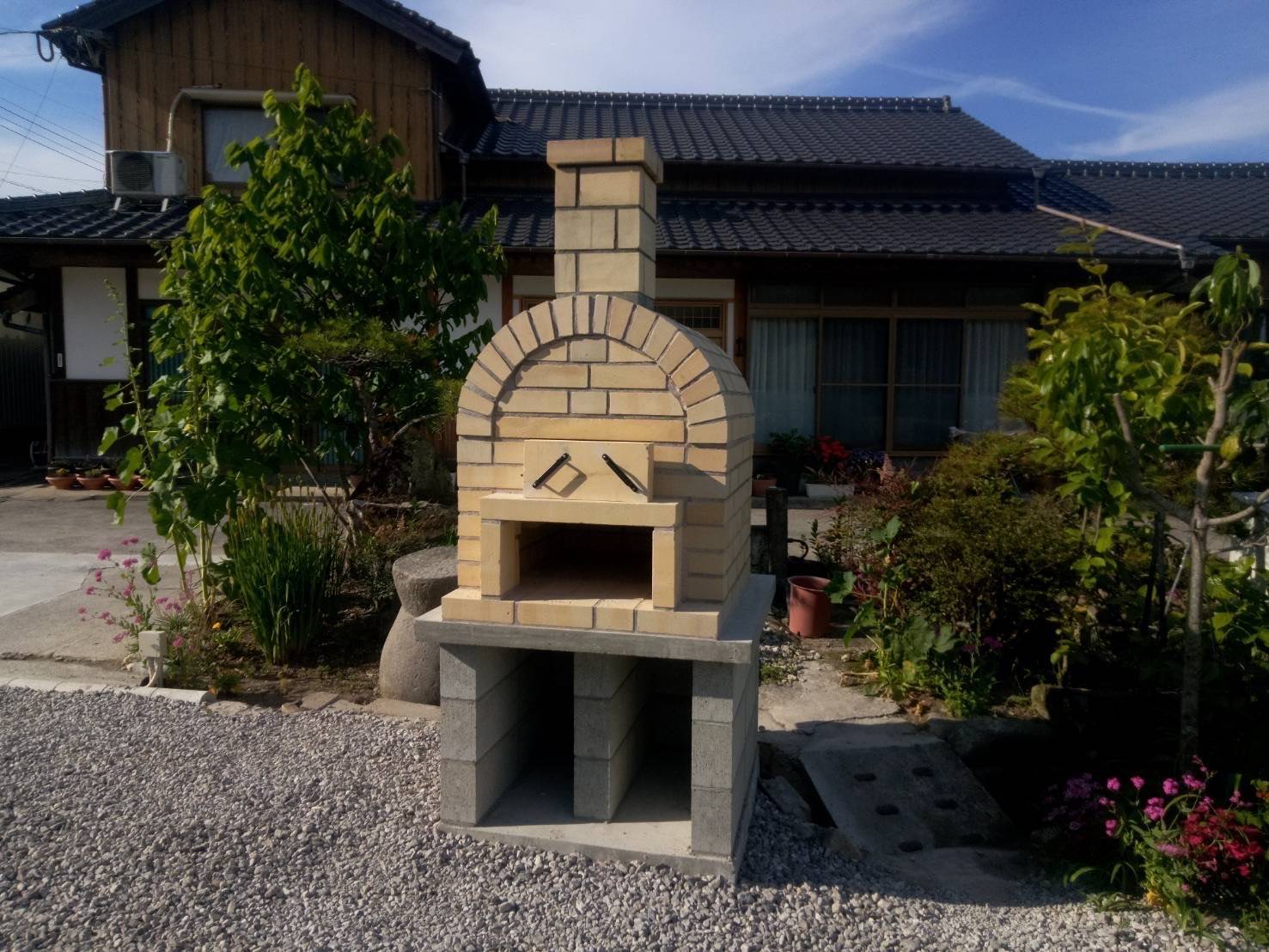 国東市の庭に設置された、オーダーメイドの屋外ピザ窯。週末のアウトドアリビングで友人たちがピザを焼いている様子。施工事例写真。